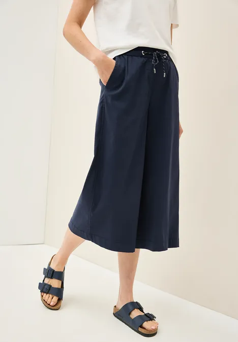 Person trägt weites blaues Culotte, weißes Shirt und Sandalen.